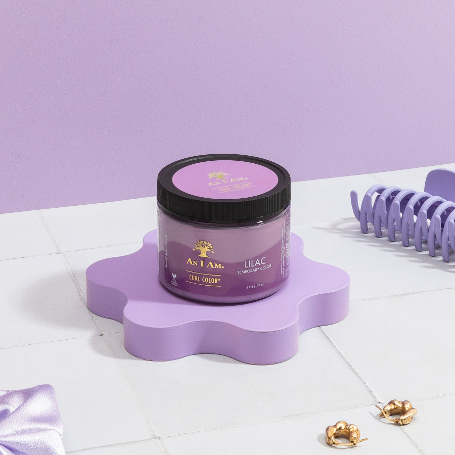عبوة من جل As I Am Curl Color Lilac للشعر المجعد موضوعة على سطح أرجواني مع مكواة تجعيد الشعر ودبابيس شعر زخرفية في الخلفية.