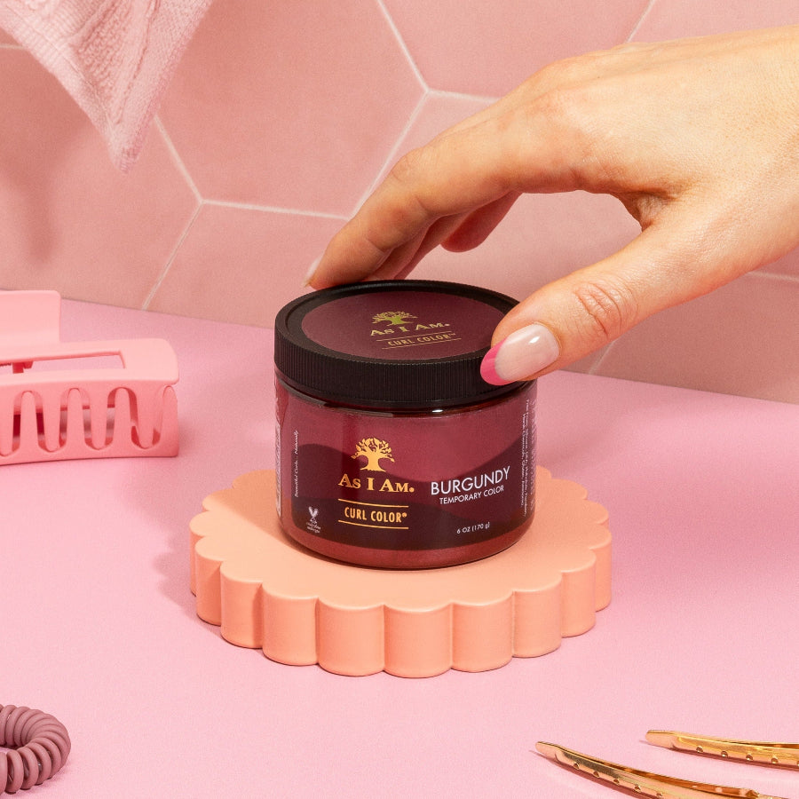 A hand holding a container of As I Am Curl Color in Burgundy, with a pink background and various hair care products.