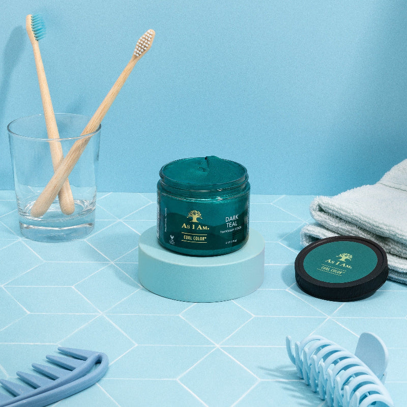 Een pot As I Am Curl Color Dark Teal Green haargel wordt naast een tandenborstel, een glas en een handdoek op een blauwe achtergrond getoond. Het product wordt gepresenteerd in een doorzichtige pot met een groene deksel en op het etiket staan de kleur en het merk vermeld.
