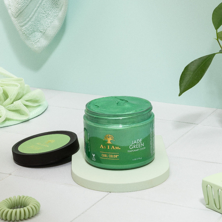 Un flacon de As I Am Curl Color en Jade Green, conçu pour les cheveux bouclés, présenté sur une surface blanche avec un couvercle vert et une plante en arrière-plan.
