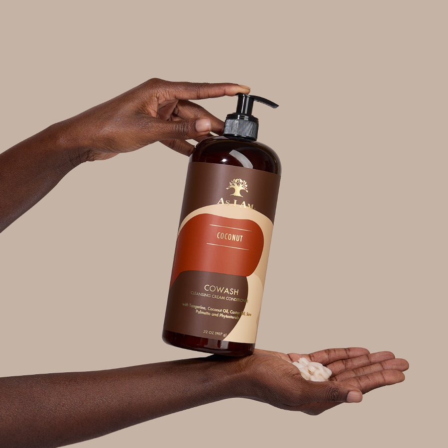 Hand met een fles As I Am Cowash Coconut conditioner met zichtbare crème op de handpalm tegen een beige achtergrond