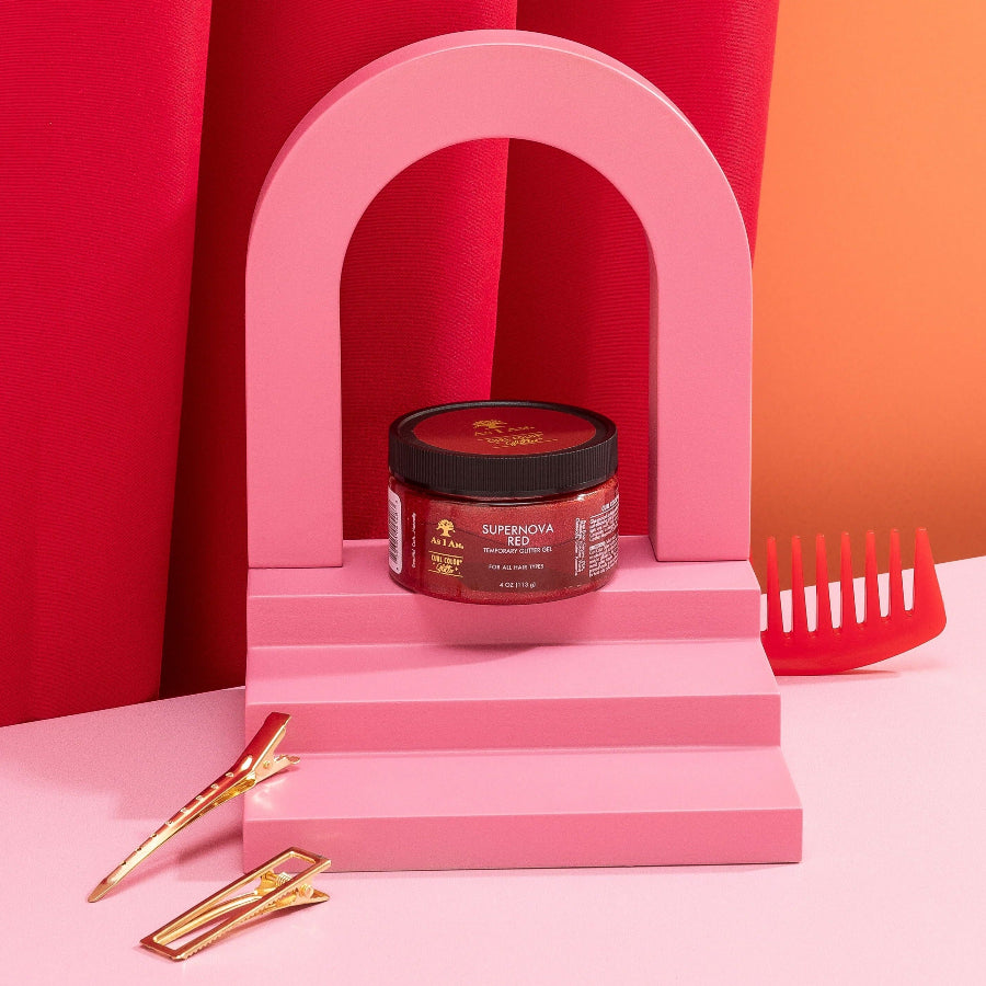 A hair color product container with a red hue, labeled 'Curl Color Glitter Supernova Red', placed on a pink stand with a brush and comb, against a backdrop of pink and orange curtains.