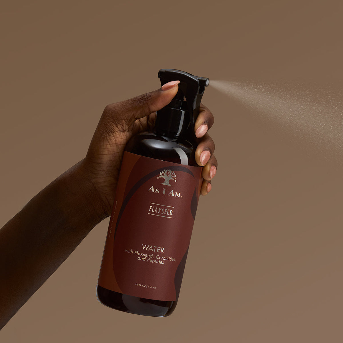 A hand holding a dark brown spray bottle labeled As I Am Flaxseed Water, spraying a fine mist against a solid warm brown background.
