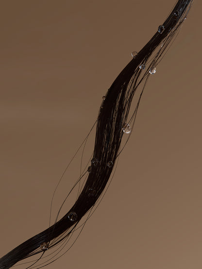 A close-up of a dark, glossy hair strand with clear water droplets hanging from it against a solid warm brown background.