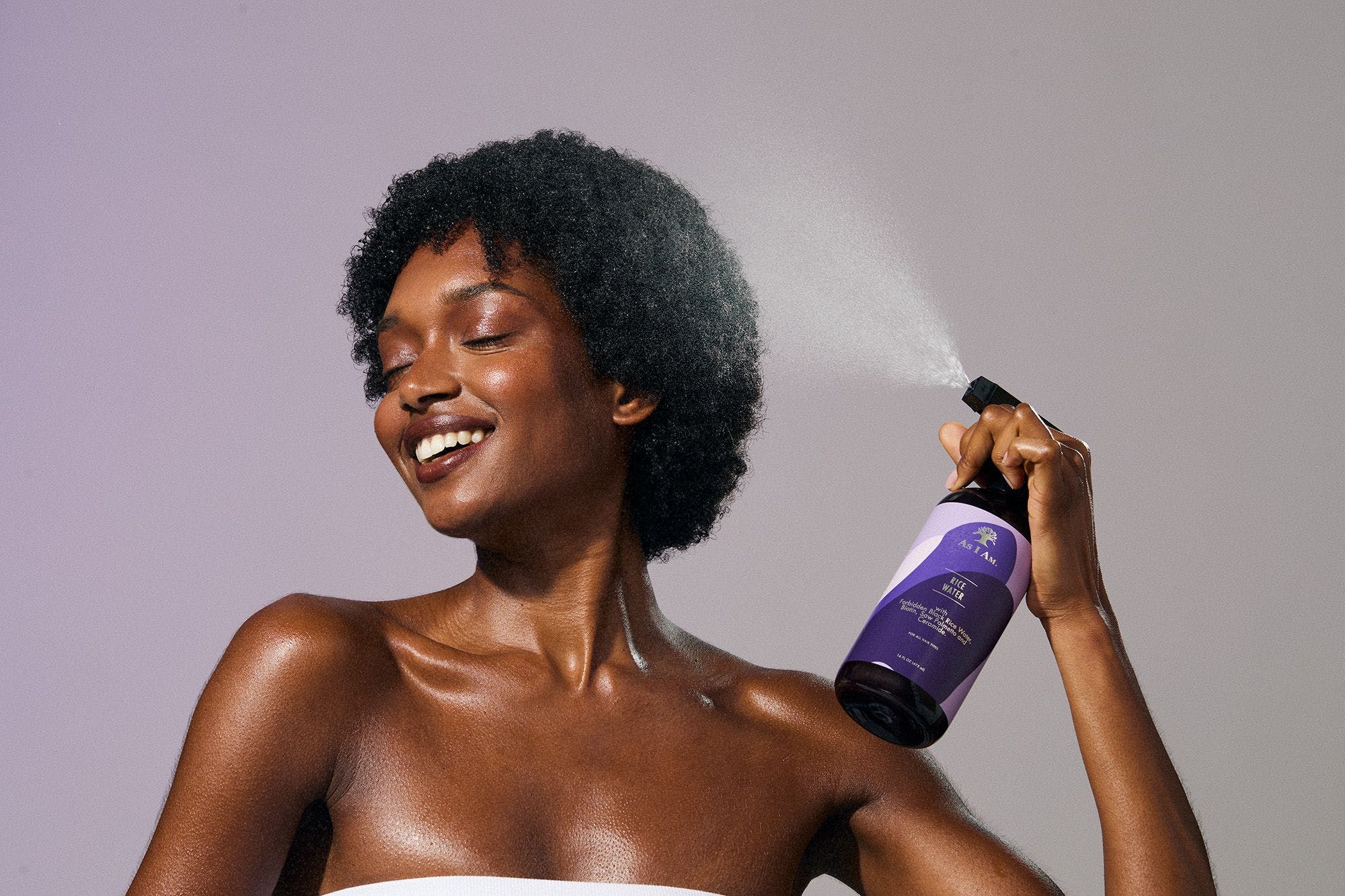 Woman applying a purple hair product spray to her hair against a plain background