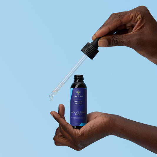 Hand holding a dropper of As I Am Dry & Itchy Scalp Care Itch Soothing Drops against a blue background