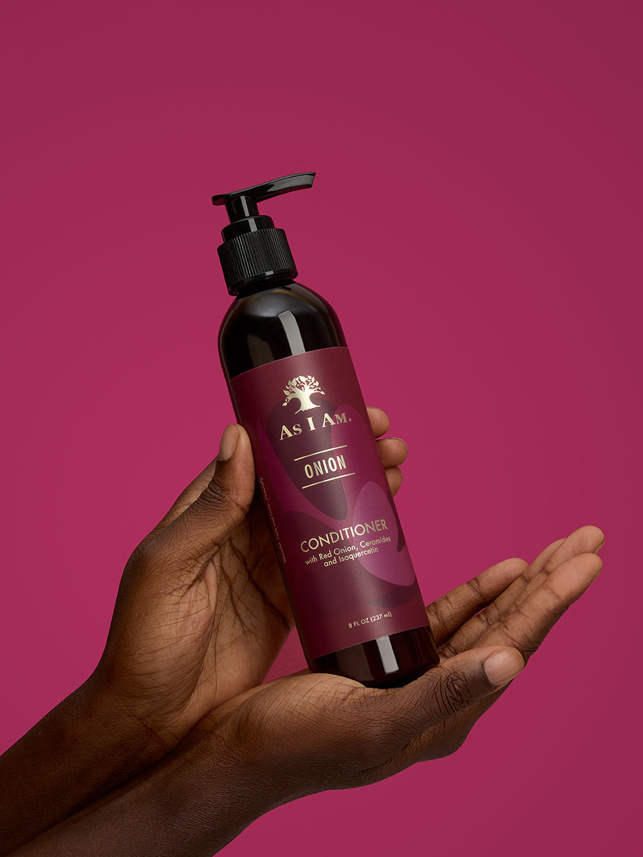 Hand, die eine Flasche As I Am Haarpflegeprodukt vor einem rosa Hintergrund hält