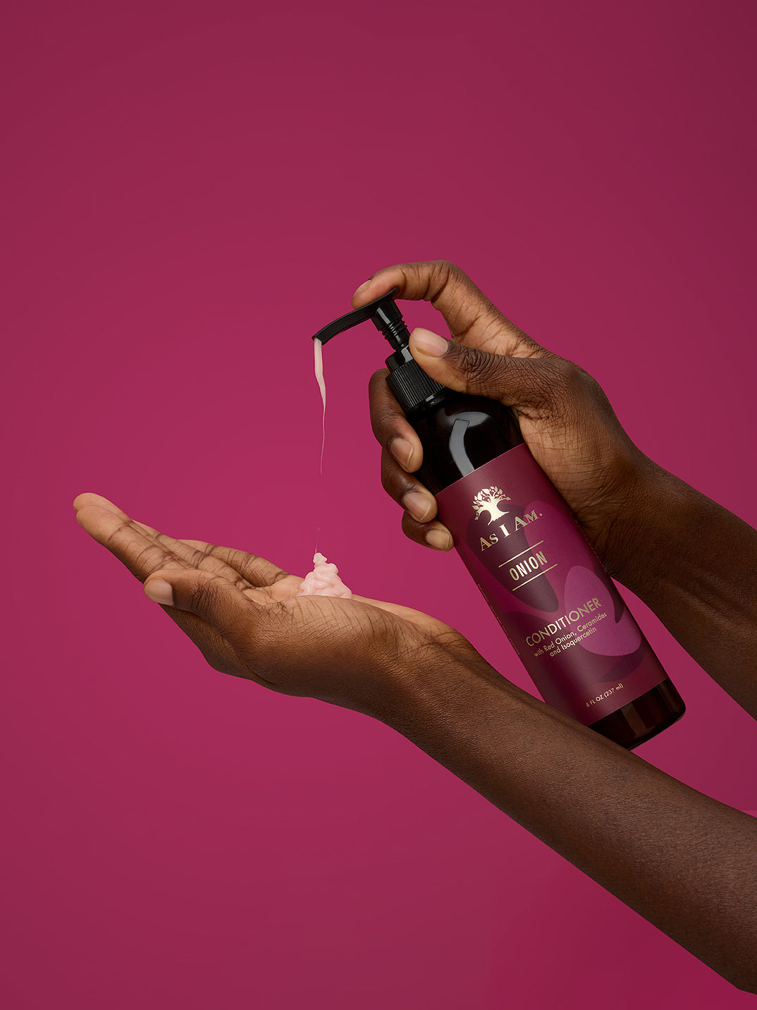Hand, die eine Flasche „as i am“-Conditioner hält, mit dem Produkt auf der Handfläche vor einem rosa Hintergrund
